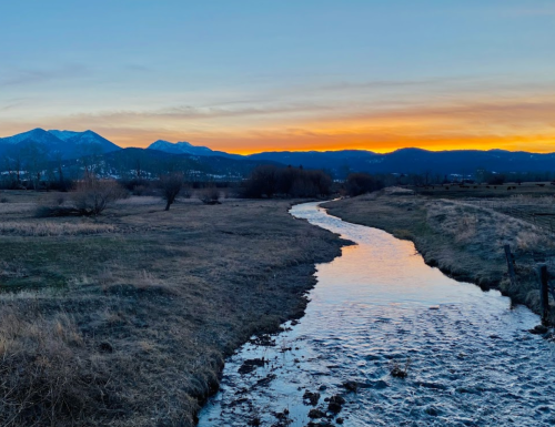 A serene landscape featuring a winding creek at sunset, with mountains in the background and a grassy field.