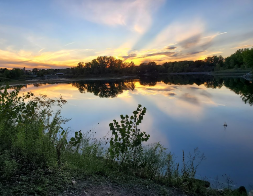 A serene lake at sunset, reflecting vibrant colors and trees along the shore, with a clear sky above.