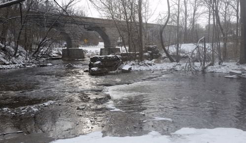 A snowy landscape featuring a creek with a stone bridge in the background, surrounded by bare trees.