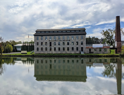 Historic stone building reflected in calm water, with a chimney and trees in the background under a cloudy sky.