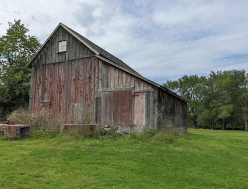 A weathered wooden barn stands in a grassy field, surrounded by trees under a cloudy sky.