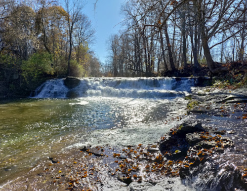 A serene waterfall cascades over rocks, surrounded by trees and autumn leaves under a clear blue sky.