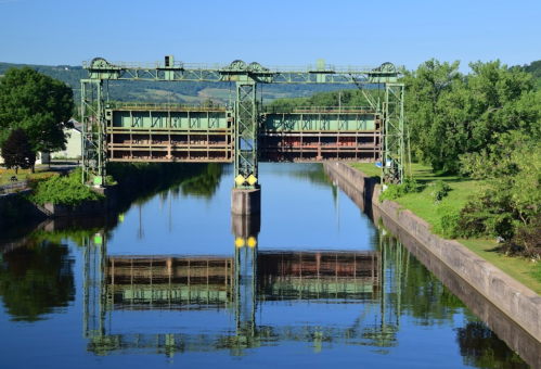 A green lift bridge spans a calm river, reflecting the structure and surrounding trees under a clear blue sky.