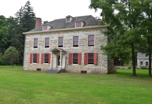 Historic stone house with red shutters, surrounded by green grass and trees.