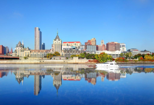 A city skyline reflects on calm water, featuring modern and historic buildings under a clear blue sky.