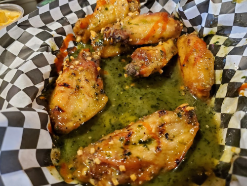 A basket of grilled chicken wings coated in a green sauce, garnished with herbs, on a checkered paper liner.