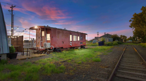 A vintage red train car converted into a cozy space, set against a colorful sunset sky and overgrown grass.