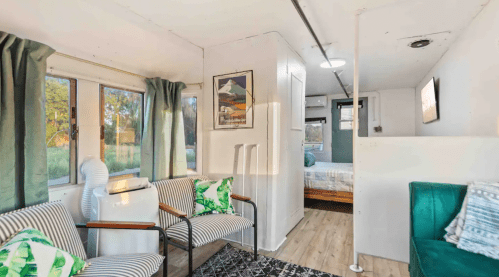 Cozy interior of a tiny home featuring seating, a bed area, and large windows with natural light.