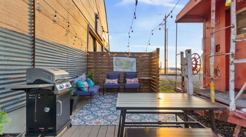 Cozy outdoor patio with seating, a grill, and string lights, beside a vintage train car at sunset.