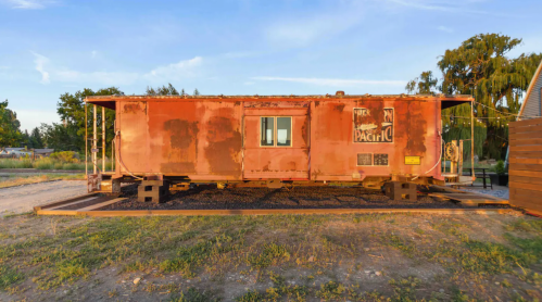A weathered red train caboose sits on a wooden platform, surrounded by grass and trees under a clear blue sky.