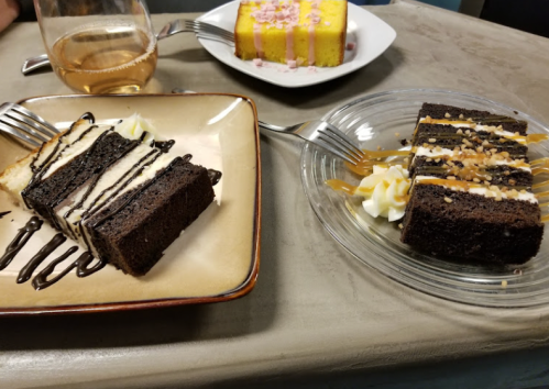 Two slices of cake on plates: one chocolate with cream and caramel, the other yellow with pink frosting. A glass of wine is nearby.