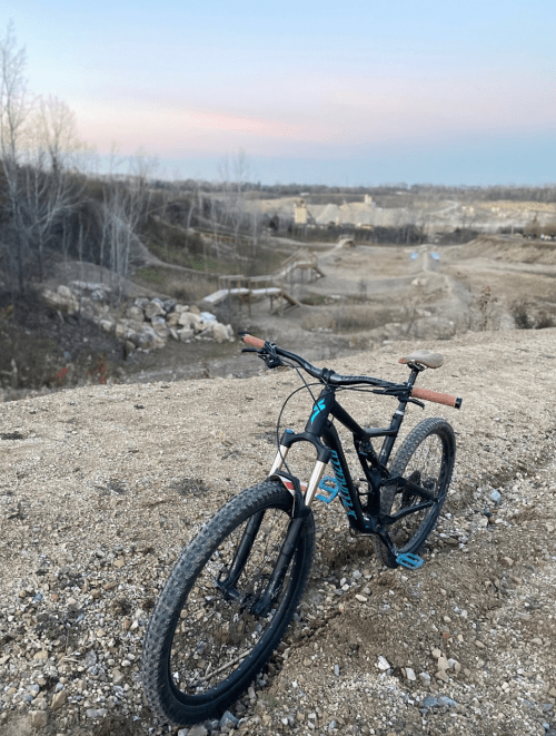 A mountain bike rests on a gravel path overlooking a scenic landscape with trees and distant hills at sunset.