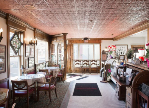 Cozy restaurant interior with ornate ceiling, wooden accents, and tables set for dining. Natural light from large windows.