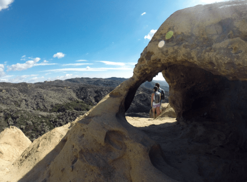 A person stands near a rock formation with a view of rolling hills and a blue sky in the background.