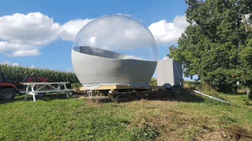 A large transparent dome structure on a wooden platform, surrounded by grass and cornfields under a blue sky.