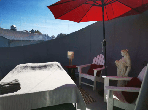 A cozy outdoor space with a massage table, two white chairs, a red umbrella, and a statue, set against a clear sky.