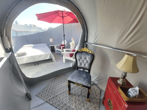 Interior of a glamping tent featuring a bed, a chair, a lamp, and a red umbrella outside. Cozy and stylish atmosphere.