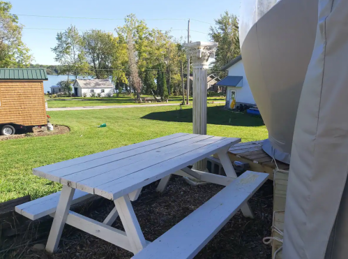 A white picnic table on a grassy area near a lake, with trees and houses in the background.
