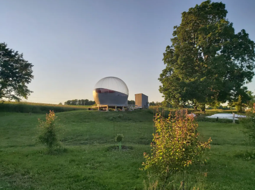 A large, spherical structure sits on a grassy hill, surrounded by trees and fields under a clear sky.