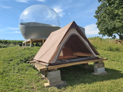 A brown tent on a wooden platform in a grassy area, with a large transparent dome structure in the background.