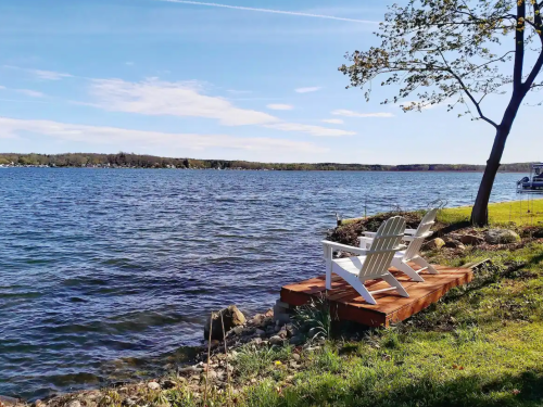 A serene lakeside view featuring two white chairs on a wooden deck, surrounded by grass and trees under a clear blue sky.
