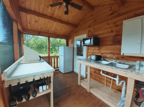 A rustic kitchen with wooden walls, featuring a fridge, microwave, sink, and cooking utensils on a countertop.