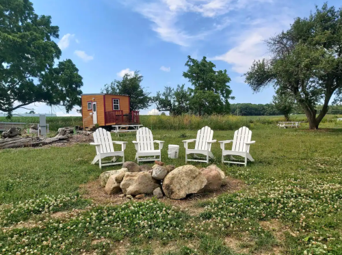 A cozy outdoor seating area with white chairs around a stone fire pit, surrounded by greenery and a small cabin.