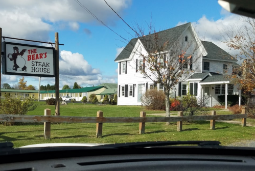 A white house with a sign for "The Bear's Steak House" in a rural setting, surrounded by green grass and trees.