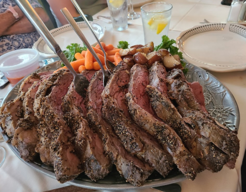 A platter of sliced prime rib with herbs, served with carrots and potatoes, alongside drinks on a dining table.
