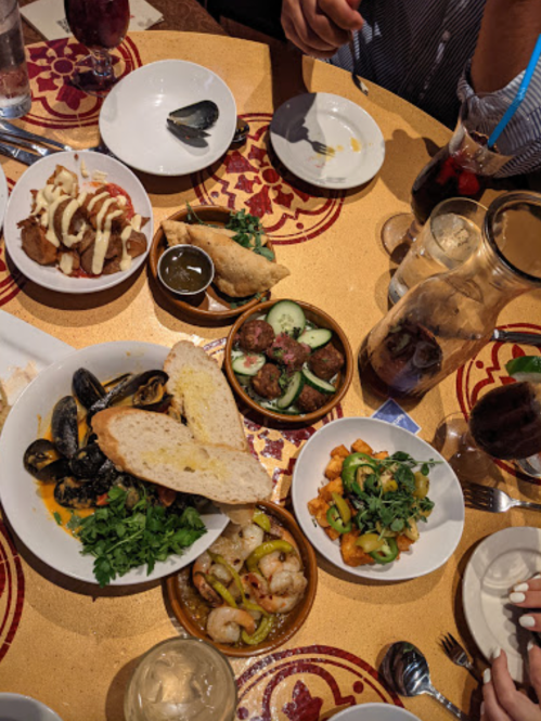 A table spread with various dishes, including seafood, bread, and appetizers, surrounded by drinks and utensils.