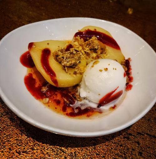 A dessert bowl with baked apples, granola topping, raspberry sauce, and a scoop of vanilla ice cream.