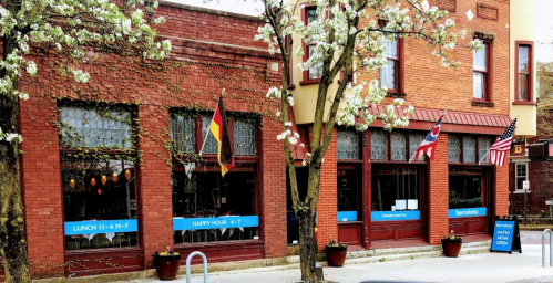 A brick building with flowering trees, featuring flags and signs for lunch, happy hour, and patio dining.