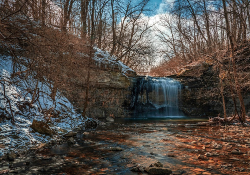 A serene waterfall cascades over rocky cliffs, surrounded by bare trees and a tranquil stream in a winter landscape.