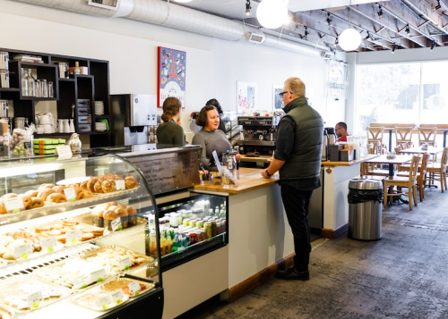 A cozy café interior with a display case of pastries, a barista serving a customer, and patrons seated at tables.