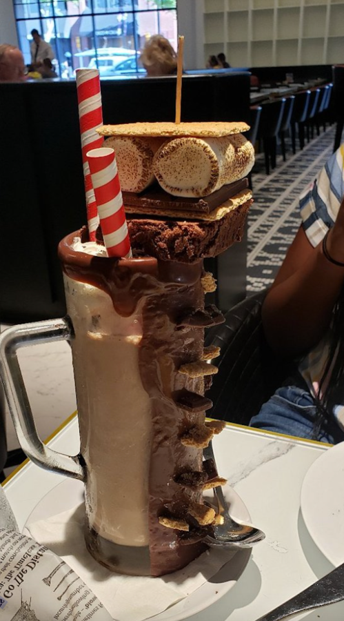 A large milkshake topped with a brownie, toasted marshmallows, and two striped straws, served in a glass mug.