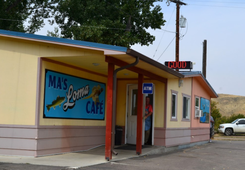 A colorful café with a sign reading "Ma's Loma Café" and an ATM, set against a clear sky and trees.