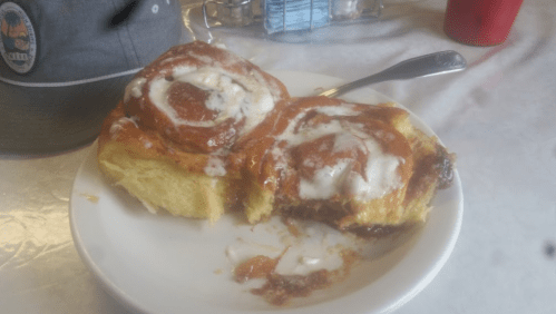 A plate with a large cinnamon roll, partially eaten, topped with icing, next to a gray hat and a glass of water.