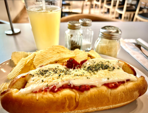 A plate with a cheesy hot dog topped with sauce, served with potato chips and a glass of lemonade.
