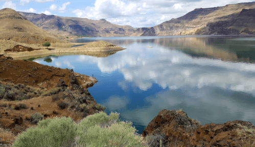 A serene lake surrounded by mountains, reflecting clouds and blue sky, with rocky shores and sparse vegetation.