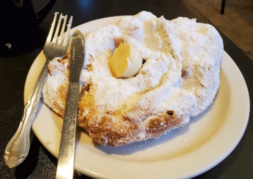 A plate with a large, powdered sugar-covered pastry topped with a pat of butter, accompanied by a fork and knife.