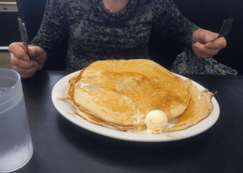 A person holds a fork and knife, ready to eat a large stack of pancakes topped with butter and syrup.