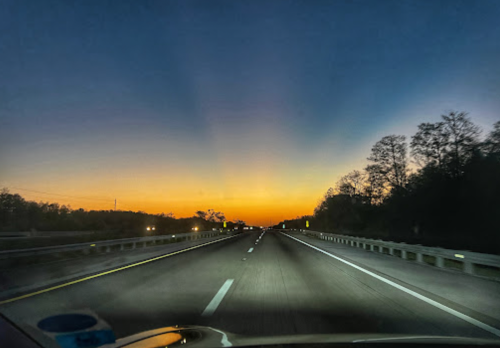 A highway at dusk with a vibrant orange and blue sunset, flanked by trees and faint headlights in the distance.