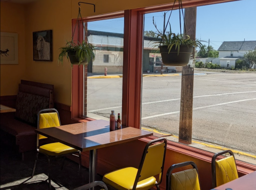 A cozy restaurant interior with yellow chairs, wooden tables, and large windows showing a sunny parking lot outside.