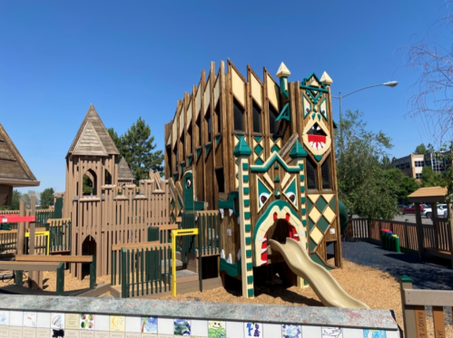 Colorful playground structure with slides, climbing areas, and a castle-like design, surrounded by trees and a sunny sky.