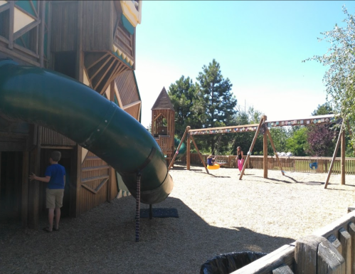 A playground featuring a large slide, swings, and children playing in a sunny outdoor setting.