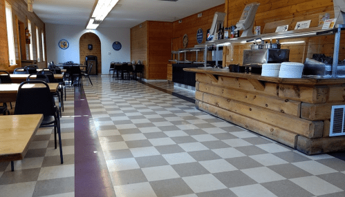 A spacious dining area with wooden walls, checkered floor, and a buffet counter. Tables and chairs are arranged neatly.