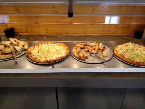 A buffet display featuring various pizzas and breadsticks on metal trays.