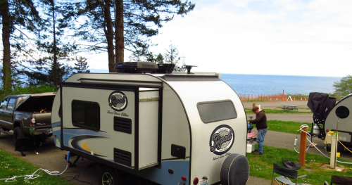 A small camper parked near the ocean, surrounded by trees and grassy area, with a truck nearby.