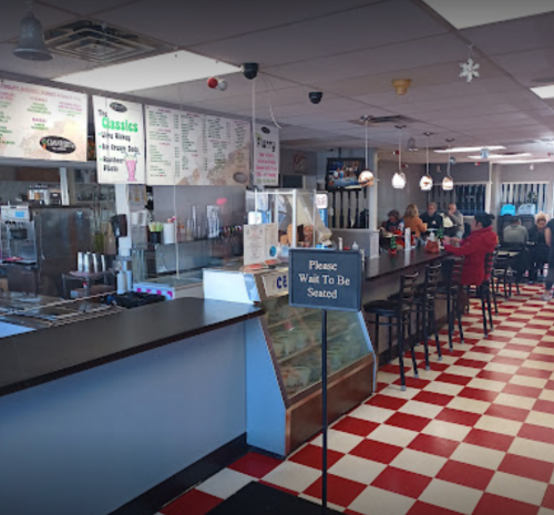 A diner interior with a checkered floor, counter, menu board, and customers seated at tables. A sign reads "Please Wait To Be Seated."