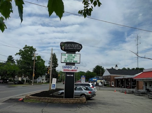 Sign for "Hawkeye's Ice Cream Parlor" with nearby businesses and a cloudy sky in the background. Cars parked in front.